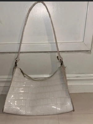 Vit axelväska i croco-mönster - Säljer en stilren vit axelväska med croco-mönstrad yta och guldfärgade metalldetaljer. Väskan har en smidig dragkedja upptill och en enkel, avlång form som passar perfekt till alla dina essentials. Materialet är syntet och remmen är bred och bekväm.