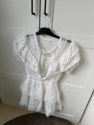 Vit volangklänning med kort ärm - Supersöt vit kortklänning med volanger och v-ringning. Klänningen har korta ärmar, markerad midja och flera lager volanger nertill. Tillverkad i ett lätt och luftigt bomullstyg, perfekt för varma dagar. Snygg detalj med knyt framtill.