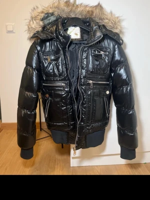 Svart glansig pufferjacka med fuskpäls - Svart pufferjacka med glansigt material, flera dragkedjefickor och huva med fuskpäls. Jackan har ribbade muddar