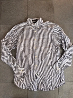 Randig skjorta från Tommy Hilfiger - Snygg skjorta från Tommy Hilfiger med blå och vita ränder. Klassisk krage, knappstängning framtill och en liten bröstficka med diskret logga. Tillverkad i mjuk bomull och har långa ärmar med manschetter. Perfekt för en clean och stilren look.