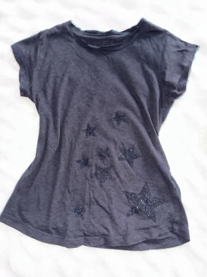  t-shirt med stjärnor från Zadig & Voltaire - Snygg mörkgrå t-shirt från Zadig & Voltaire med glittriga stjärnor framtill. Lite Tecken på användning och köpt second hand, knappt några stenas fattas.  Den ska vara lite ”sliten” uppe vid halsen för att modellen ser ut så. Storlek XS-S