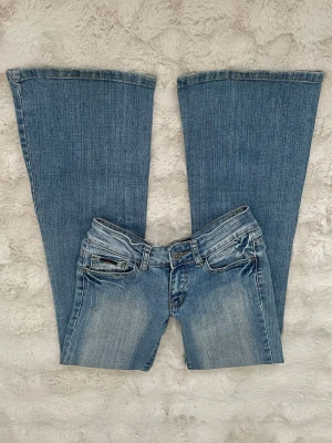 Blå bootcut jeans med låg midja - Snygga blå bootcut jeans med låg midja och klassisk femficksdesign. Jeansen har en ljus tvätt och subtila slitningar framtill för en cool vintagekänsla. Perfekta för dig som gillar en avslappnad och trendig stil.