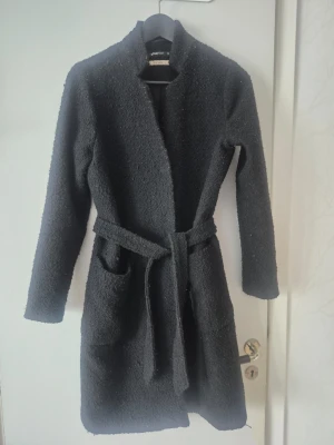 Svart kappa med bälte - Stilren svart kappa med bälte i midjan och stora fickor framtill. Kappan har lång ärm och rak passform, perfekt för kyligare dagar. Materialet är mjukt och har en lätt textur som ger en modern känsla.