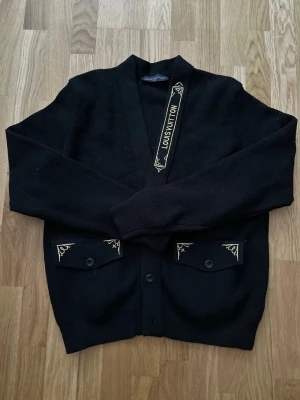 Svart cardigan från Louis Vuitton - Svart cardigan med knappar från Louis Vuitton. Tröjan är i nytt skicka och har aldrig använts då den är för liten för mig. Stoleken är XL men sitter som L. Tröjan har två fickor framtill med dekorativa broderier i gult på ficklocken och en snygg detalj med Louis Vuitton-logga på insidan. Perfekt för en stilren och exklusiv look.