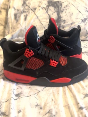Svarta och röda Air Jordan 4 sneakers - Snygga Air Jordan 4 sneakers i svart och rött med klassisk Jumpman-logga på hälen. Skorna har meshpaneler, röda detaljer och svart mocka på ovandelen. Tjock sula med röd och svart färgkombination. Perfekta för dig som gillar streetwear och basketstil.