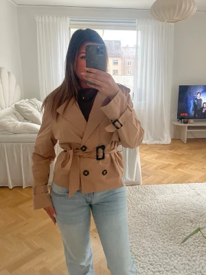 Kort beige trenchcoat med bälte - Snygg kort trenchcoat i beige med dubbelknäppning, bälte i midjan och svarta spänndetaljer. Klassisk krage och justerbara ärmslut. Perfekt för en trendig och stilren look.