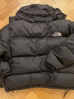 The North Face 700 svart puffer jacket - Säljer en svart pufferjacka från The North Face med hög krage och justerbar dragsko nedtill. Jackan har broderad logga på bröstet och breda, vadderade paneler som ger en riktigt fet siluett. Perfekt för kalla vinterdagar och riktigt street.
