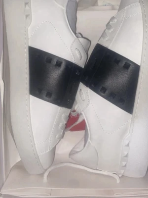 Valentino rock stud open - Dessa Valentino Garavani-sneakers har en elegant vit design med en svart banddetalj. De verkar vara i utmärkt skick utan synliga tecken på slitage.  ⭐️ Skick: 9/10 🎨 Färg: Vit och svart 📦 Snabb och noggrann leverans