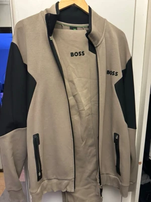 Beige BOSS set - Nypris ca: 3000kr.                                              Snyggt loungewear-set från BOSS i beige med svarta detaljer på ärmar och sidor. Jackan har dragkedjor på fickorna och BOSS-logga på bröstet. Matchande byxor ingår. Perfekt för en avslappnad och stilren look hemma.     Storlek L på byxor och XXL på tröja.