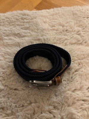 Mörkblått flätat bälte med läderdetaljer - Snyggt mörkblått flätat bälte med bruna läderdetaljer och silverspänne. Perfekt accessoar för att lyfta din outfit med en klassisk touch. Bältet har en stilren design och passar till många olika looks.