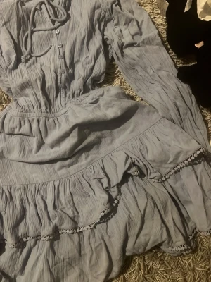 Ljusblå volangklänning med knappar - Supersöt ljusblå klänning med volanger och spetsdetaljer. Klänningen har långa ärmar med resår vid ärmslut, knappar framtill och en knytning vid halsen. Tillverkad i ett mjukt, krinklat bomullstyg som ger en avslappnad känsla. Jätte fin till sommaren men kommer inte till användning finns lite defekter som missfärgning som inte syns men kolla sista bilden 