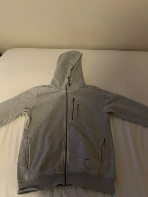 Grå hoodie från Sail Racing - Snygg grå hoodie från Sail Racing med dragkedja framtill, huva och tre fickor med dragkedja. Mjuk insida och diskret logga på bröstet. Perfekt för en avslappnad och sportig stil.