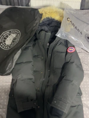 Svart dunjacka från Canada Goose - Säljer en svart dunjacka från Canada Goose med klassisk patch på ärmen och avtagbar huva med fuskpäls. Jackan är lång, har flera fickor och stängs med dragkedja och knappar. Perfekt för kalla vinterdagar och riktigt varm.