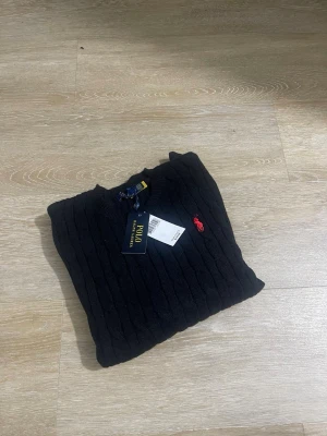 Svart kabelstickad tröja Polo Ralph Lauren - Svart kabelstickad tröja från Polo Ralph Lauren med rund halsringning och klassisk röd broderad logga på bröstet. Tröjan har en tidlös design och är tillverkad i mjukt material som passar perfekt till många olika outfits.