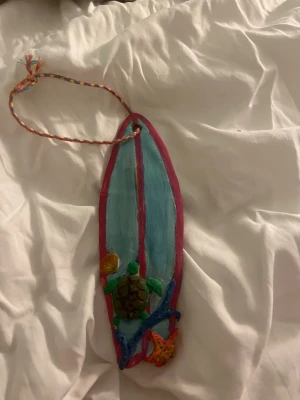 Dekorativ surfbräda med sköldpadda - Färgglad dekoration i form av en surfbräda med blå och rosa färger, prydd med en liten sköldpadda, snäcka och korallmotiv. Hängs upp med ett flätat snöre. Perfekt som accessoar till väskan eller rummet för en strandig vibe.