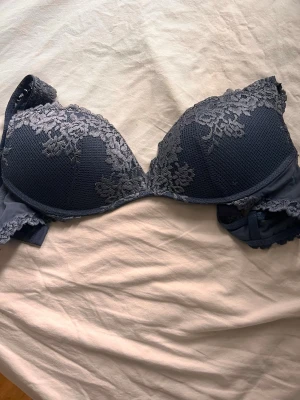 Mörkblå intimissimi push-up bh med spetsdetaljer - Snygg mörkblå push-up bh med vadderade kupor och dekorativa spetsdetaljer i grått över kuporna och längs kanterna. Bh:n har justerbara axelband och hakknäppning bak. Materialet är en mix av mjuk textil och spets som ger en lyxig känsla.
