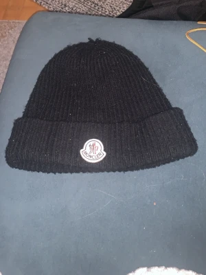 Svart ribbstickad mössa från Moncler - Säljer en svart ribbstickad mössa från Moncler med uppvikt kant och Moncler-logga framtill. Klassisk och stilren design som passar perfekt till höst och vinter. Tillverkad i mjukt material som håller dig varm.