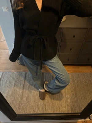 Svart omlottkofta med knytband - Säljer en svart omlottkofta med knytband i midjan. Koftan har långa ärmar och en djup v-rigning. Enkel och stilren design som passar till det mesta.