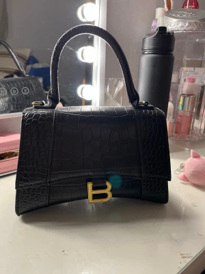 Svart handväska i croc-mönster - Säljer en svart handväska med croc-embossat mönster och guldfärgad B-detalj framtill. Väskan har ett stadigt handtag och en elegant, strukturerad form. Perfekt för dig som vill ha en lyxig accessoar med statement-känsla.