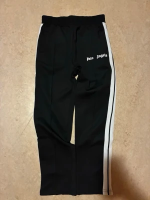 Palm Angels svarta mjukisbyxor - Svarta mjukisbyxor från Palm Angels med vita sidoränder och logga på vänster sida. Byxorna har resår i midjan och dragkedjor vid bensluten för extra stil och funktion. Tillverkade i ett mjukt och följsamt material som passar perfekt för en avslappnad look.