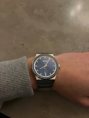 Blå BOSS klocka med silverlänk - En äkta stilren klocka från BOSS med blå urtavla och datumvisning. Klockan har en klassisk rund form och en robust silverfärgad länk i metall. Perfekt accessoar för dig som gillar en clean och modern look.