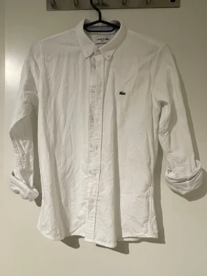Vit skjorta från Lacoste - Klassisk vit skjorta från Lacoste med button-down krage och den ikoniska krokodilloggan på bröstet. Skjortan har långa ärmar och knäppning framtill. Perfekt till både jeans och chinos för en clean look. Storlek 164-170cm.