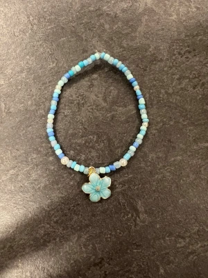 Blått pärlarmband med blomberlock Storlek M  - Ett armband med pärlor i olika blå nyanser och vitt. Armbandet har ett hänge i form av en ljusblå blomma med gulddetaljer. 
