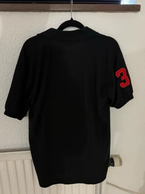 Svart pikétröja från Polo Ralph Lauren - Svart pikétröja med klassisk krage från Polo Ralph Lauren. Stor röd broderad logga framtill och röd siffra 3 på ärmen. Perfekt för en sportig och stilren look.
