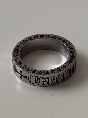 Silverring från Chrome Hearts - Snygg silverfärgad ring från Chrome Hearts med ingraverad svart text och korsdetaljer runt hela ringen. Ringen har en bred, platt design och är tillverkad i metall med en cool, industriell känsla. Perfekt för dig som gillar statement-smycken och streetwear. Ringen är oanvänd (ny). Kan gå ner i pris vid snabb affär. Skriv vid minsta lilla fundering!