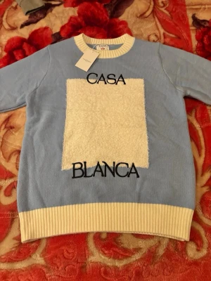 Blå stickad tröja från Casablanca - Ljusblå stickad tröja från Casablanca med vit ribbad mudd vid hals, ärmslut och nederkant. Framsidan har en stor vit, fluffig ruta och svart text 'CASA BLANCA'. Tröjan har en avslappnad passform och är långärmad. Perfekt för dig som gillar trendiga stickade plagg.