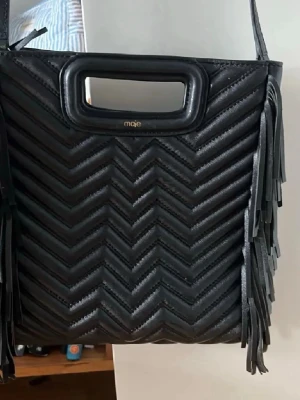Maje väska  - Snygg svart handväska från Maje i quiltat konstläder med chevronmönster. Väskan har fransar på sidorna och ett inbyggt handtag upptill. Perfekt accessoar för att lyfta din outfit med en cool och trendig touch.