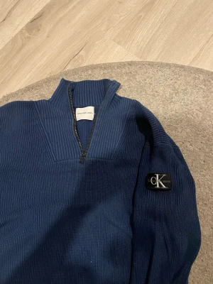 Blå stickad tröja från Calvin Klein - Mörkblå ribbstickad tröja från Calvin Klein Jeans med halv dragkedja och hög krage. Tröjan har en snygg CK-logga på ärmen och är tillverkad i ett mjukt, tjockt material som håller värmen. Perfekt för dig som gillar stilrena och klassiska plagg.