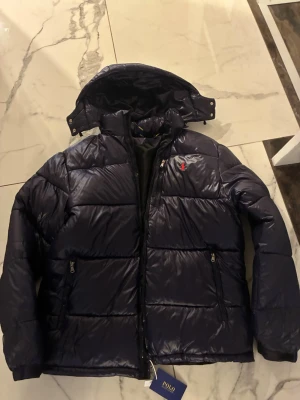 Mörkblå dunjacka från Polo Ralph Lauren - Snygg mörkblå dunjacka från Polo Ralph Lauren med huva och dragkedja. Jackan har två framfickor med dragkedja och klassisk Polo-logga på bröstet. Perfekt för kyliga dagar och riktigt stilren look. Storlek M men är stor i storleken så skulle säga köp om du har L. Säljer då den var för stor för mig