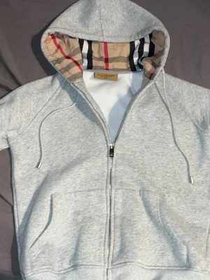 Grå zip-hoodie från Burberry - Snygg grå hoodie med dragkedja från Burberry. Klassisk huva fodrad med det ikoniska rutmönstret i beige, svart och rött. Mjukt material, dragsko i huvan och två fickor framtill. Perfekt för en stilren och avslappnad look.