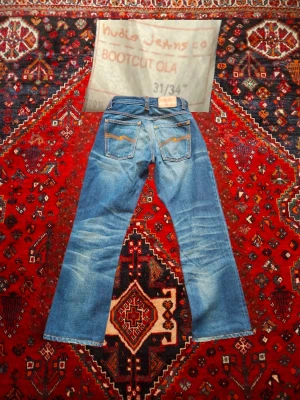 Nudie Jeans Bootcut Ola  - Bootcut Ola jeans från Nudie Jeans i klassisk blå denim med snygga slitningar och kontrastsömmar. Bakfickorna har Nudies ikoniska broderi och passformen är bootcut med normal midja. Perfekta för dig som gillar en avslappnad men ändå stilren look. Mindre I storlek enligt mig, be om mått om så önskas 