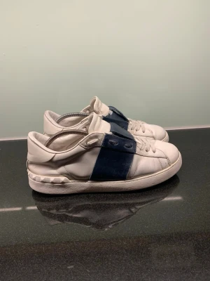 Valentino open - Snygga vita sneakers från Valentino med en bred marinblå rem över mitten och nitar på hälen. Skorna är låga och har klassisk snörning. Tillverkade i läder med en stilren och modern design som sticker ut. Storlek 43 i mycket bra skick. Skickar 1-2 efter köp!