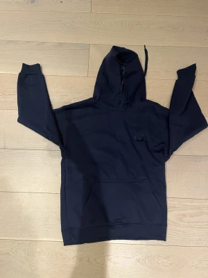 Mörkblå hoodie med huva - Snygg mörkblå hoodie med stor känguruficka framtill och broderad logga på bröstet. Klassisk modell med ribbade muddar vid ärmslut och nederkant. Tillverkad i mjukt bomullsmaterial och har justerbar dragsko i huvan.