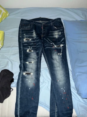 Jeans Dsquared2 - Säljer ett par mörkblå jeans från Dsquared2 med slitningar, hål och färgstänk för en riktigt edgy look. Jeansen har fem fickor, raka ben och tydliga tvättade detaljer framtill och baktill. Perfekta för dig som gillar streetwear och statement-plagg.