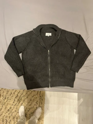 Grå stickad ziptröja Maison Margiela - Snygg grå stickad ziptröja från Maison Margiela med grov ribbstickning och hög krage. Tröjan har hel dragkedja framtill och är perfekt för lager-på-lager. Klassisk och stilren design som funkar till mycket. Storlek M och använda ett  tag. Men väldigt bra skick och inga defekter.