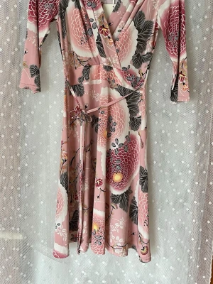 Blommig omlottklänning Stockholms Klänningsfabrik - Supersnygg blommig omlottklänning från Stockholms Klänningsfabrik i rosa, vinrött och grått. Klänningen har trekvartsärm, knytband i midjan och ett vackert blommönster. Perfekt för dig som gillar feminina och färgglada plagg. Djur och rökfritt 