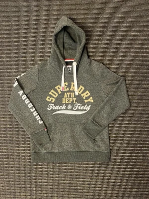 Superdry hoodie - Mörkgrön hoodie från Superdry med stor gul och vit logga på bröstet och tryck längs armen. Finns inga täcken av andväning. Perfekt för en skönt och avslappnad stil.  Hör av dig vid minsta fundering 💯💫