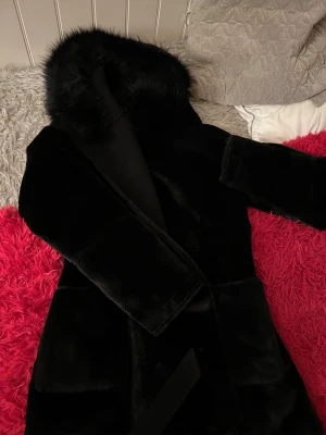 Svart salsa jeans pälskappa  - Snygg svart pälskappa med fluffig huva och bälte i midjan. Jackan har långa ärmar och en mjuk, lyxig känsla. Perfekt för dig som vill ha en varm och stilren look under kalla dagar. Jackan har även praktiska fickor och en elegant siluett. Nypris 3599kr