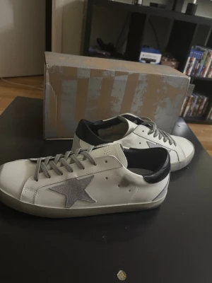 Vita golden goose med grå stjärna  - Snygga vita sneakers från GGDB med grå stjärna på sidan, svarta detaljer vid hälen och grå snörning. Skorna har en klassisk låg profil och platt sula, perfekta för en trendig streetstyle-look. Hör av dig vid minsta lilla fundering