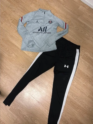 PSG träningströja & Under Armour byxor - Snygg ljusgrå långärmad träningströja från PSG med svarta och röda detaljer samt svart logga. Matchas med svarta träningsbyxor från Under Armour med vita sidor och logga på låret. Perfekt för träning eller sport.