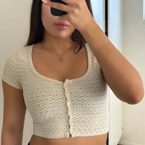 Beige croppad blus med knappar - Söt croppad blus i beige med korta ärmar och dekorativa knappar framtill. Toppen har ett hålmönster och är ribbad, vilket ger en trendig och luftig känsla. Perfekt för varmare dagar och enkel att styla med olika outfits.
