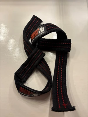 Svarta lifting straps Gorilla Wear - Svarta liftingstraps från Gorilla Wear med röda sömmar. Remmarna är tillverkade i slitstarkt tyg och har en klassisk design med logotyp på etiketten. Perfekta för styrketräning och gym.