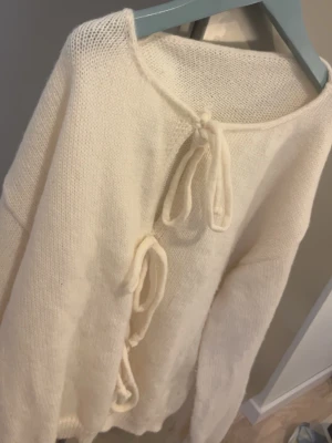 Krämvit stickad tröja med knyt - Mysig krämvit stickad tröja med tre dekorativa knyt framtill. Tröjan har rund halsringning och långa ärmar. Materialet ser ut att vara mjukt och fluffigt, perfekt för kyligare dagar. Enkel men trendig design som passar till mycket.