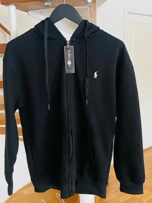 Svart hoodie från Polo Ralph Lauren - Snygg svart hoodie från Polo Ralph Lauren med dragkedja framtill och klassisk vit broderad logga på bröstet. Tillverkad i mjukt bomullsmaterial för en skön känsla. Hoddien är även oanvänd för den passad ej mig, bara testad.