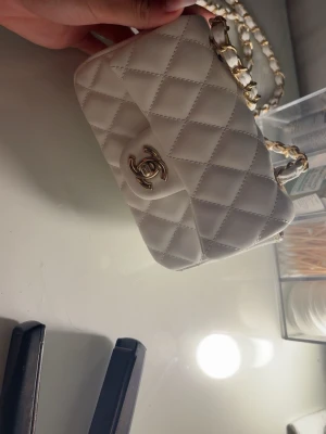 Vit quiltad Chanel handväska - Supersnygg vit quiltad handväska från Chanel med klassiskt guldigt CC-spänne framtill. Kedjerem med insytt vitt läder ger en lyxig vibe. Perfekt storlek för det viktigaste och ikonisk design som aldrig går ur tiden.
