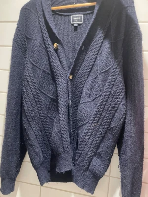 Mörkblå stickad kofta från Bakers - Mysig mörkblå stickad kofta från Bakers med kabelstickat mönster och två guldfärgade knappar framtill. Koftan har långa ärmar och ribbade muddar vid ärmslut och nederkant. Perfekt för lager på lager och chill stil. L/XL